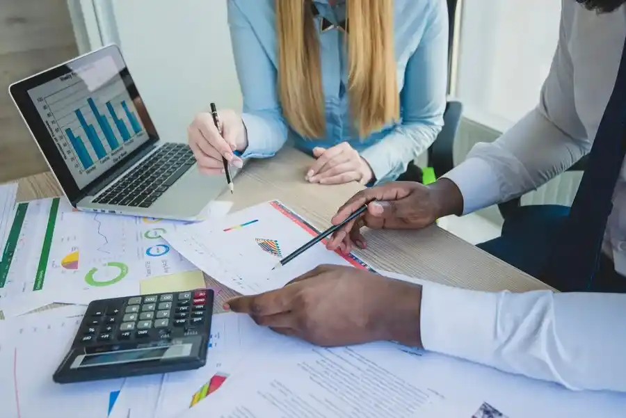 A imagem exibe um grupo de profissionais em uma reunião, analisando documentos e gráficos de desempenho financeiro. Eles estão envolvidos em atividades de planejamento e análise de dados, o que é relevante para o contexto do artigo "ICMS-ST – Novo percentual de IVA-ST para o estado de São Paulo". A presença de um laptop com gráficos de barras e de uma calculadora indica que eles podem estar examinando os impactos financeiros de uma nova alíquota de imposto, como o ICMS-ST (Imposto sobre Circulação de Mercadorias e Serviços de Transição), no estado de São Paulo. A discussão e a anotação de informações sugerem que eles estão preparando-se para ajustar suas operações financeiras e estratégias de negócios de acordo com as novas taxas de imposto, o que é crucial para a gestão financeira e a conformidade fiscal das empresas.