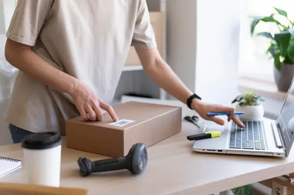 A imagem mostra uma pessoa preparando um pacote para envio, com um laptop ao lado, simbolizando a integração entre o varejo físico e o online. No contexto do artigo, isso reflete a tendência de unificar as lojas físicas e virtuais, para fornecer uma experiência de compra mais integrada e conveniente para os clientes, além de aproveitar as vantagens de ambos os canais para aumentar as vendas e a fidelidade do cliente.