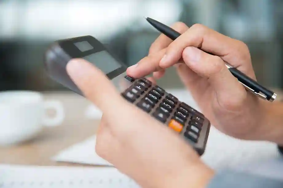 Close-up das mãos de um profissional de contabilidade utilizando uma calculadora financeira e segurando uma caneta sobre documentos de impostos. A imagem representa o cálculo de tributos estaduais e a importância da emissão da GNRE para a conformidade fiscal no e-commerce e logística interestadual. Fundo suavemente desfocado com uma xícara de café, sugerindo um ambiente de trabalho administrativo e financeiro.