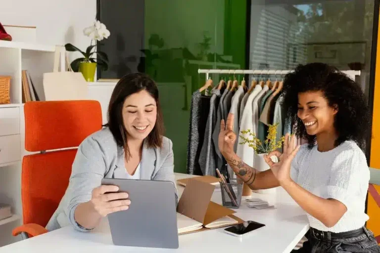 A imagem mostra duas mulheres em um ambiente de trabalho ou loja, interagindo de forma ativa com dispositivos digitais. A mulher à esquerda está olhando para um tablet com um sorriso, enquanto a mulher à direita está dando palmas, sugerindo uma celebração ou aprovação. O ambiente é agradável e informal, com roupas penduradas no fundo, o que pode indicar uma loja de varejo ou um espaço de trabalho relacionado ao comércio.