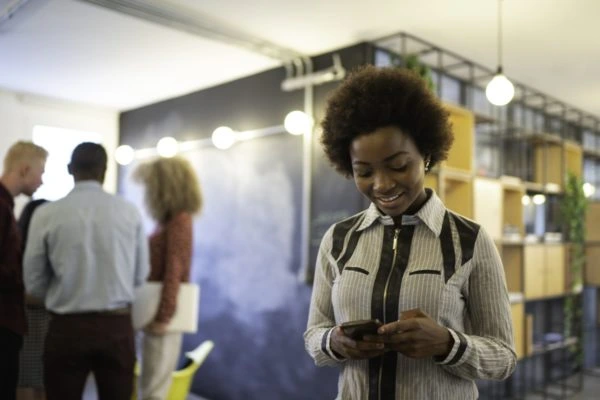 Mulher sorridente usando smartphone em um ambiente de exposição comercial, com outros profissionais ao fundo. Representa o uso de aplicativos B2B para impulsionar relacionamentos e vendas entre empresas.
