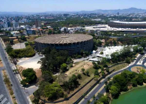 A imagem mostra uma vista aérea da região de Belo Horizonte, com destaque para um grande estádio de futebol circular, provavelmente o Mineirão. Ao redor do estádio, é possível ver a paisagem urbana da cidade, com edifícios, áreas verdes e outras infraestruturas. Essa imagem é muito relevante para o artigo "ONCLICK tem nova unidade em Belo Horizonte", pois ela ilustra a localização e o contexto da nova unidade da empresa na capital mineira. O estádio Mineirão, um dos principais cartões-postais da cidade, serve como um ponto de referência visual para situar a nova unidade da ONCLICK no cenário da cidade.