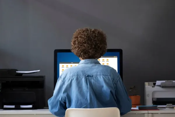 A imagem mostra uma pessoa sentada em uma mesa, olhando para um computador que está mostrando uma interface de gerenciamento de arquivos, provavelmente um sistema de backoffice. O ambiente sugere um local de trabalho, talvez o escritório de uma empresa ou o lar de uma pessoa que trabalha remotamente. No contexto do artigo "Conheça os mecanismos de backoffice que levam o seu e-commerce a gerar valor", a imagem pode ser usada para ilustrar a importância do backoffice em operações de e-commerce. O backoffice é o coração do gerenciamento de uma loja online, lidando com tarefas críticas como processamento de pedidos, gerenciamento de estoque, contabilidade, marketing e suporte ao cliente.