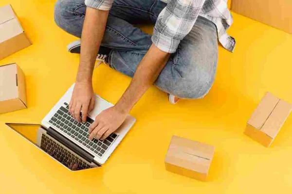 Homem sentado no chão com um notebook e caixas ao seu redor, simbolizando gerenciamento de vendas online. Representa a ideia de criar ou gerenciar um negócio digital de forma independente e flexível.