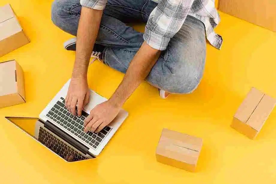 Homem sentado no chão com um notebook e caixas ao seu redor, simbolizando gerenciamento de vendas online. Representa a ideia de criar ou gerenciar um negócio digital de forma independente e flexível.