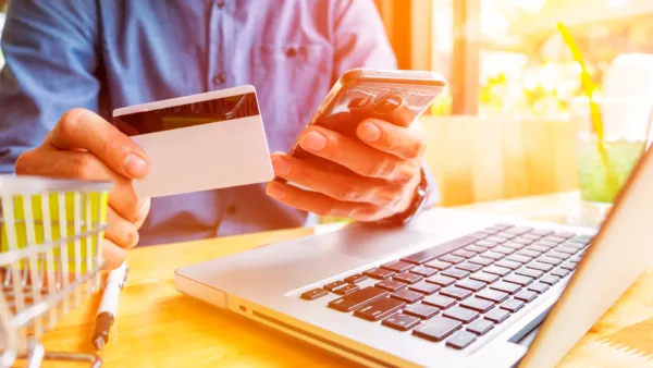 A imagem exibe uma pessoa realizando uma transação online com um cartão de crédito, segurando o cartão na mão esquerda e tomando uma foto do cartão com um smartphone na mão direita. O laptop está em frente a ele, o que sugere que ele está no processo de fazer uma compra ou check-out no e-commerce. No contexto do material "Conheça 5 aplicações essenciais para a gestão do seu e-commerce!", a imagem pode ser usada para ilustrar a importância das ferramentas digitais e aplicativos na gestão e operação de um negócio de e-commerce.