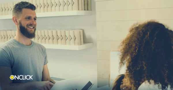 Homem sorridente segurando um tablet enquanto atende uma cliente em um ambiente profissional. A imagem ilustra a tendência de usar tecnologia para melhorar a experiência do cliente, um dos temas do artigo sobre tendências de customer sucess. Logo da ONCLICK no canto inferior esquerdo.