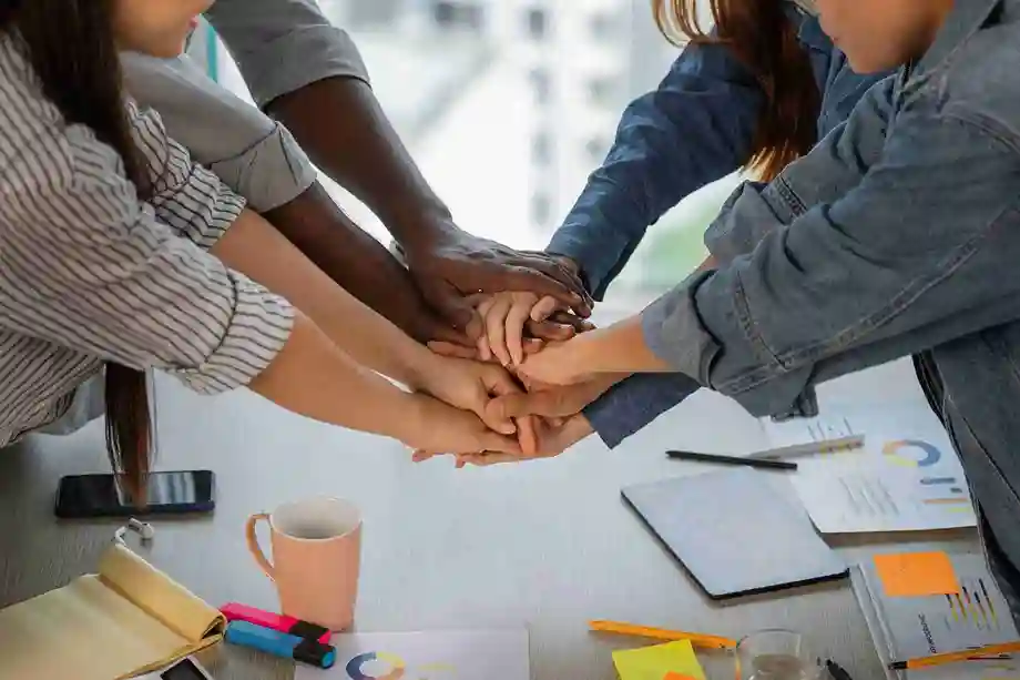 A imagem exibe uma equipe de colaboradores unindo mãos, refletindo o espírito de equipe e a colaboração. No contexto do artigo, isso destaca a importância de selecionar e treinar talentos adequados para trabalhar em uma loja, enfatizando a necessidade de um time engajado e capacitado para impulsionar o sucesso do negócio e fornecer uma ótima experiência ao cliente.