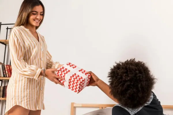 A imagem mostra uma mulher entregando um presente envolvido em papel com um padrão de ziguez para uma criança, que está sentada e estendendo a mão para receber o presente. A mulher está sorrindo e parece satisfeita com a entrega, enquanto a criança está ansiosa e feliz por receber o presente. No contexto do artigo "Dia das crianças: aumente as vendas do seu e-commerce nesta data", a imagem pode ser usada para ilustrar a importância de aproveitar datas comemorativas, como o Dia das Crianças, para impulsionar as vendas do e-commerce.