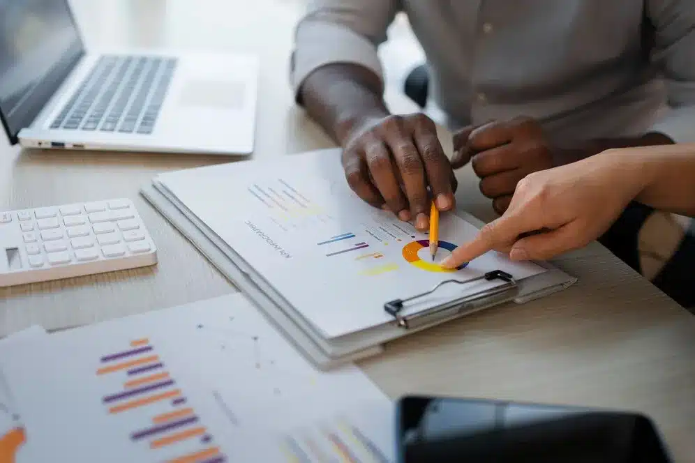A imagem mostra uma pessoa analisando relatórios financeiros ou de negócios em uma folha de papel, enquanto outro indivíduo aponta para a folha. O ambiente é um escritório com um laptop e um teclado, o que indica um ambiente de trabalho corporativo. A pessoa que está analisando os relatórios está usando uma caneta para apontar detalhes nos gráficos. No contexto do artigo "Como os relatórios inteligentes facilitam a gestão do seu negócio", a imagem pode ser usada para ilustrar como a tecnologia e os relatórios inteligentes podem simplificar e melhorar a análise de dados para tomada de decisão.