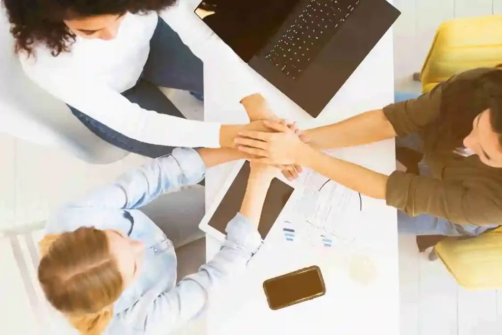 A imagem mostra uma equipe de trabalho unindo mãos em torno de uma mesa com dispositivos digitais, simbolizando a colaboração e o engajamento em um ambiente varejista. No contexto do artigo, isso reflete a importância de equilibrar a gestão de equipes e das máquinas, destacando a necessidade de integração entre tecnologia e pessoas para melhorar a eficiência e a satisfação do cliente no varejo.