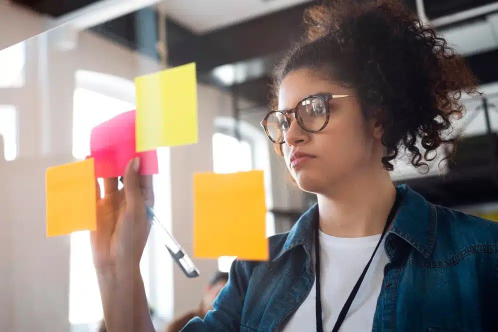 A imagem mostra uma mulher concentrada, usando óculos e analisando notas adesivas coloridas em uma parede. Isso sugere que o artigo "Saiba o que são Metodologias Ágeis e as suas vantagens" provavelmente abordará os seguintes tópicos. O objetivo do artigo deve ser apresentar uma visão geral sobre as Metodologias Ágeis, suas características e os benefícios que elas podem trazer para organizações que buscam maior flexibilidade e eficiência em seus projetos.