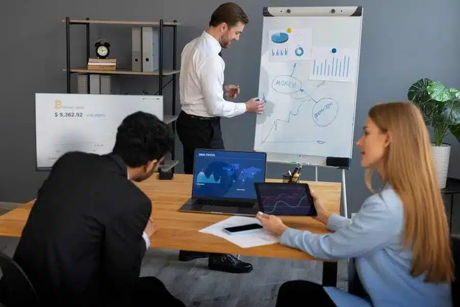 A imagem mostra uma reunião de equipe em um ambiente de escritório. Há um homem apresentando informações em um quadro branco, enquanto outros membros da equipe estão sentados em torno de uma mesa, analisando gráficos e dados em seus laptops. Essa imagem é relevante para o artigo "Os desafios da Gestão de Projetos e como lidar com eles", pois ela retrata uma situação típica de uma reunião de acompanhamento de projetos, onde a equipe está envolvida na análise de indicadores e no planejamento de ações.