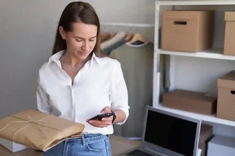 A imagem mostra uma mulher em um ambiente de armazenamento ou de estoque, olhando para um smartphone com uma expressão de concentração. À sua frente, há uma mesa com caixas de papelão e um laptop, o que sugere que ela está lidando com tarefas relacionadas ao gerenciamento de estoque ou ao comércio eletrônico. No contexto do artigo "Da loja física ao marketplace: como ter um controle de estoque eficiente", a imagem pode ser usada para ilustrar a importância de uma gestão eficaz do estoque para as empresas que operam em diferentes canais de comércio, como lojas físicas e marketplaces.