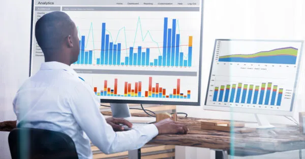Homem analisando gráficos e dados em um computador, com um monitor grande mostrando gráficos de colunas e linhas. Representa o trabalho de análise de dados, business intelligence ou monitoramento de desempenho. O ambiente é um escritório com uma mesa de madeira e um ambiente de trabalho organizado.