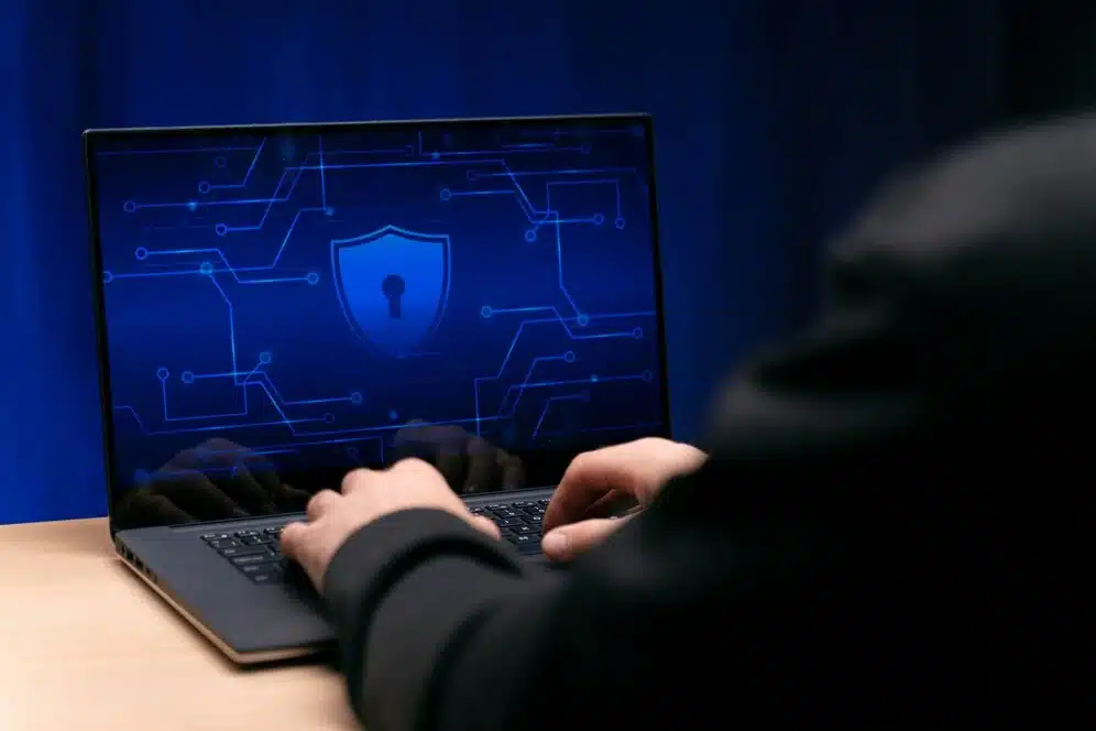 A imagem apresenta uma tela de computador com um símbolo de escudo e circuitos eletrônicos ao fundo, indicando um tema relacionado à segurança da informação. Isso se alinha com o título do artigo "Saiba como aplicar a segurança da informação no seu negócio". Portanto, a imagem reforça o foco do artigo em orientar os leitores sobre como aplicar efetivamente a segurança da informação para proteger seus negócios de riscos digitais.