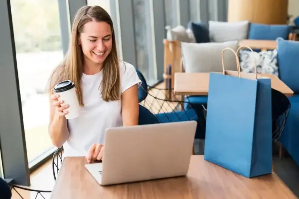 A imagem mostra uma mulher sorridente trabalhando em um laptop em um ambiente doméstico. Ela está segurando uma xícara de café e parece estar fazendo compras online, com sacolas de compras ao seu redor. Essa cena pode ser relevante para o artigo "Panorama do e-commerce: como se preparar para o segundo semestre de 2021", pois retrata uma situação típica de comércio eletrônico, com uma consumidora realizando suas atividades de compra em um ambiente confortável.