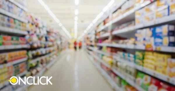 Corredor de supermercado com prateleiras cheias de produtos, iluminado por luzes fluorescentes. O logo da ONCLICK aparece no canto inferior esquerdo. A imagem tem um efeito de desfoque suave, criando uma sensação de amplitude e organização no ambiente comercial.