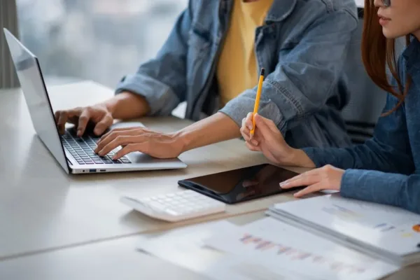 A imagem exibe uma cena de trabalho colaborativo, com duas pessoas sentadas em uma mesa, trabalhando juntas. Uma delas está digitando em um laptop, enquanto a outra, com uma caneta em mãos, parece estar analisando ou anotando informações de um documento. No meio da mesa, há um smartphone, um teclado sem fio e mais alguns papéis, sugerindo um ambiente de trabalho ativo e produtivo. No contexto do artigo "Consultoria completa, especializada e adaptada com a ONCLICK", a imagem pode ser usada para representar a dinâmica de trabalho em equipe e a importância da consultoria especializada na otimização de processos e na tomada de decisões informadas.