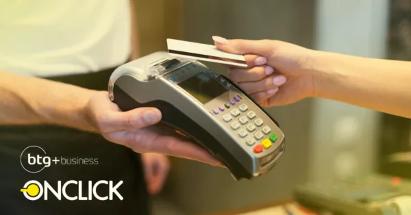 A imagem mostra uma pessoa realizando um pagamento com cartão de crédito em um terminal de POS (Point of Sale). A mão que segura o terminal está prestes a processar a transação, enquanto a outra mão segura o cartão que será usado para pagar. No canto inferior esquerdo da imagem, há logos de "b+g+business" e "ONCLICK", indicando a associação entre as duas empresas. No contexto do artigo "Conheça a parceria ONCLICK & BTG+ business", a imagem provavelmente ilustra a funcionalidade e o objetivo da parceria entre as duas empresas, que é fornecer soluções de pagamento inovadoras e eficientes para os negócios.