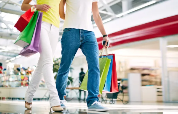 A imagem mostra dois consumidores caminhando em um centro comercial, cada um carregando várias sacolas de compras de diferentes lojas. A cena reflete a experiência do cliente no varejo físico, onde os indivíduos realizam compras em várias lojas e interagem com diferentes marcas. No contexto do material "Jornada do cliente: como desenhá-la para o seu negócio", a imagem pode ser usada para ilustrar a importância de entender e mapear a jornada do cliente, que envolve todos os pontos de contato e interações que um cliente tem com uma empresa ou marca ao longo do tempo.