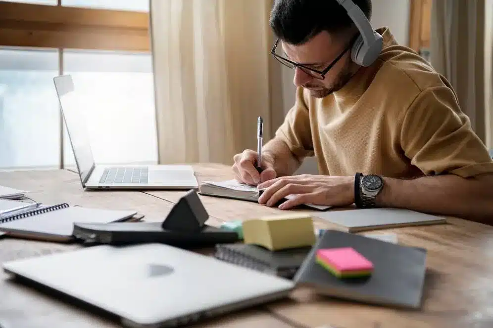 A imagem mostra um homem trabalhando em seu computador portátil em um ambiente de escritório. Ele está usando fones de ouvido e parece estar concentrado em suas atividades. Há diversos materiais de escritório espalhados sobre a mesa, como blocos de notas, canetas e um smartphone, o que sugere que ele está envolvido em algum tipo de trabalho ou projeto.