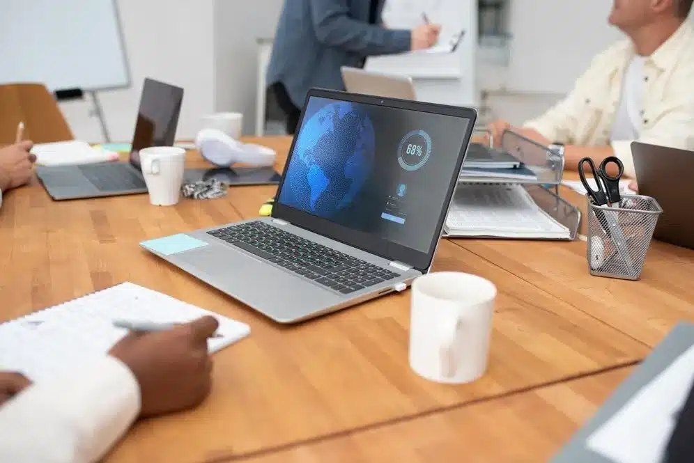 A imagem mostra uma reunião de trabalho onde várias pessoas estão sentadas em torno de uma mesa, com notebooks e documentos. O notebook no centro da imagem exibe uma tela com um gráfico global e um percentual, o que sugere a análise de dados ou a monitoração de performance global. Os participantes estão envolvidos em atividades de trabalho, como anotar em papéis e interagir com dispositivos digitais.