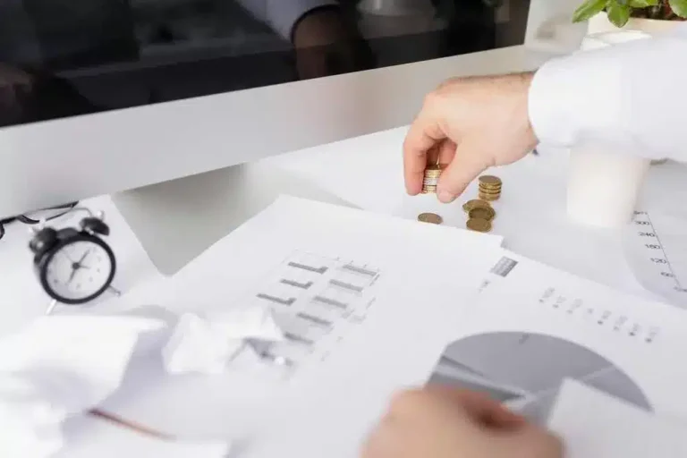 A imagem mostra uma mão segurando moedas sobre uma pilha de papéis com gráficos e informações financeiras. Isso sugere um contexto relacionado a finanças e análise de dados. Essa imagem é relevante para o artigo "O que é Open Finance e qual o impacto no e-commerce?" pois ela representa aspectos relacionados à gestão financeira e à análise de dados, que são temas importantes no contexto do Open Finance e seu impacto no comércio eletrônico.