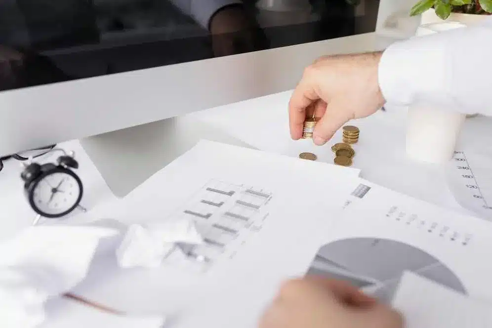 A imagem mostra uma mão segurando moedas sobre uma pilha de papéis com gráficos e informações financeiras. Isso sugere um contexto relacionado a finanças e análise de dados. Essa imagem é relevante para o artigo "O que é Open Finance e qual o impacto no e-commerce?" pois ela representa aspectos relacionados à gestão financeira e à análise de dados, que são temas importantes no contexto do Open Finance e seu impacto no comércio eletrônico.