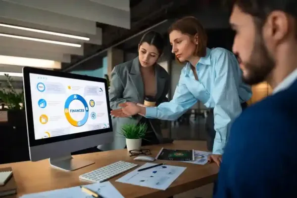 A imagem mostra uma reunião de trabalho onde três indivíduos estão examinando um monitor que exibe gráficos e informações financeiras. A pessoa no centro está apontando para o monitor, possivelmente discutindo os dados com os colegas, enquanto um laptop, papéis e um tablet estão dispostos na mesa, sugerindo uma sessão de análise de performance ou planejamento.