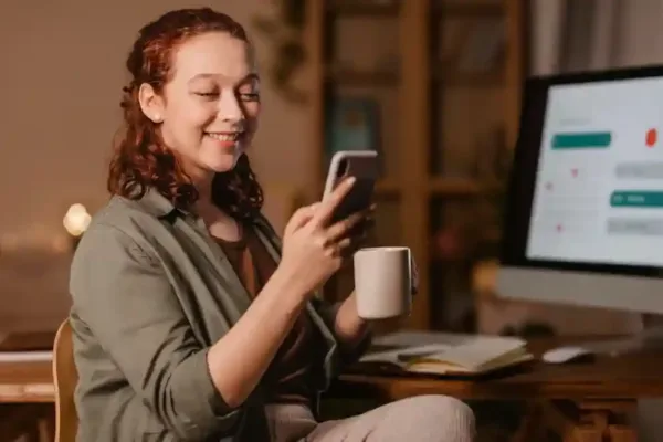A imagem mostra uma mulher sorrindo e olhando para um smartphone, que segura com uma mão, enquanto a outra mão segura um caneca. Ela está sentada em um ambiente de trabalho, com um computador e documentos à sua frente. A cena sugere que ela está interagindo de forma positiva com seu smartphone, possivelmente obtendo informações úteis ou gerenciando seu negócio.
