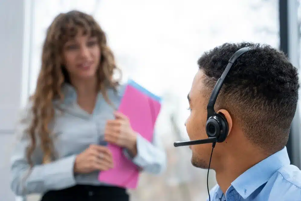 A imagem mostra uma mulher segurando pastas rosa e um homem usando um fone de ouvido com microfone, indicando uma situação de atendimento ao cliente ou de treinamento. Isso se alinha com o título do artigo "Novo decreto do SAC", que provavelmente aborda atualizações ou mudanças nas regulamentações do Serviço de Atendimento ao Consumidor (SAC).