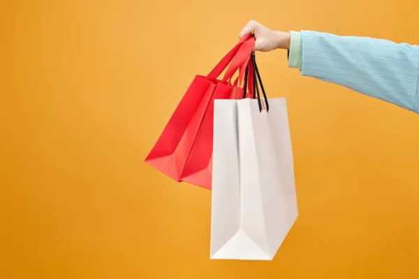Mão segurando duas sacolas de compras, uma vermelha e uma branca, em um fundo amarelo. Representa a experiência do cliente e a importância de adicionar valor ao seu negócio através de experiências de compra satisfatórias e personalizadas.