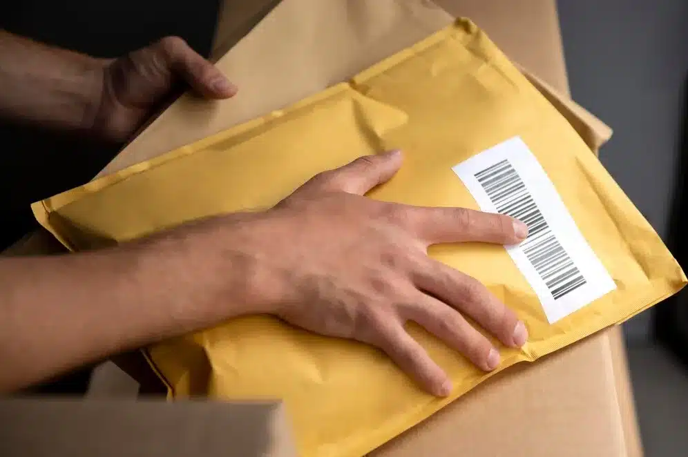 A imagem mostra uma pessoa segurando uma embalagem de correio com uma etiqueta de rastreamento. A embalagem é amarela e está sobre um fundo de caixas de papelão, o que sugere um ambiente de logística ou distribuição. No contexto do artigo "Qual é a relação entre e-commerce e Mercado Livre?", a imagem pode ser usada para ilustrar o processo de entrega de produtos adquiridos através de plataformas de e-commerce, como o Mercado Livre. Mercado Livre é uma plataforma popular para o comércio eletrônico no Brasil, que permite aos usuários comprar e vender produtos online