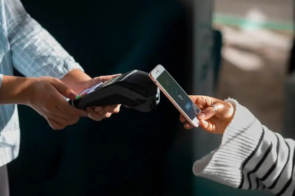 A imagem mostra uma pessoa realizando uma transação de pagamento utilizando o PIX, um sistema de pagamentos instantâneos desenvolvido pelo Banco Central do Brasil. É possível ver as mãos da pessoa segurando um smartphone e um terminal de pagamento, indicando que a transação está sendo feita em um ambiente de comércio eletrônico.