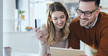 A imagem mostra um casal em um ambiente familiar, provavelmente em casa, olhando para um smartphone com expressões de satisfação. A mulher está segurando o smartphone e parece estar compartilhando uma experiência agradável com o homem, que está ao seu lado e também está olhando para o dispositivo com um sorriso. Eles estão em frente de um laptop, o que pode estar exibindo informações relevantes para a atividade que estão realizando no smartphone. No contexto do material "Guia completo para otimizar a experiência do cliente no seu e-commerce", a imagem pode ser usada para ilustrar a importância de uma experiência do cliente positiva e personalizada.