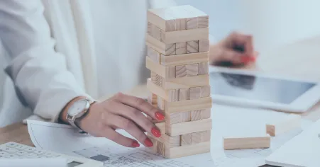 A imagem mostra uma pessoa construindo uma estrutura de blocos de madeira, um por um, em uma mesa. Em segundo plano, há um laptop e papéis, o que sugere um ambiente de trabalho ou de estudo. A pessoa está focada na tarefa, o que indica uma atenção meticulosa e cuidadosa. No contexto do material "Gestão de risco: um guia para aplicar no seu negócio", a imagem pode ser usada para ilustrar a importância de construir uma gestão de risco sólida e estratégica para o negócio, assim como os blocos de madeira precisam ser cuidadosamente colocados para construir uma estrutura estável.
