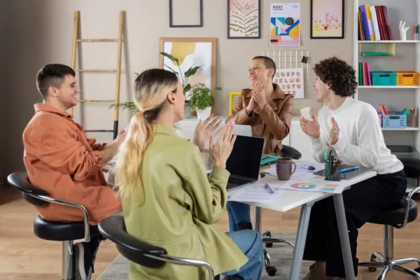A imagem mostra um grupo de quatro pessoas sentadas em volta de uma mesa em um ambiente de escritório, aparentemente celebrando um momento de sucesso ou aprovação. Eles estão apertando as mãos, sorrisos no rosto, o que indica um ambiente de trabalho positivo e de equipe unida. A mesa é cheia de papéis, um laptop e outros materiais de trabalho, sugerindo que eles estão em meio a uma reunião ou discussão de projeto. No contexto do artigo "Qual o papel da Cultura Corporativa na Retenção de Trabalhadores do Varejo", a imagem pode ser usada para ilustrar a importância de uma cultura corporativa saudável e inclusiva na retenção de talentos no varejo