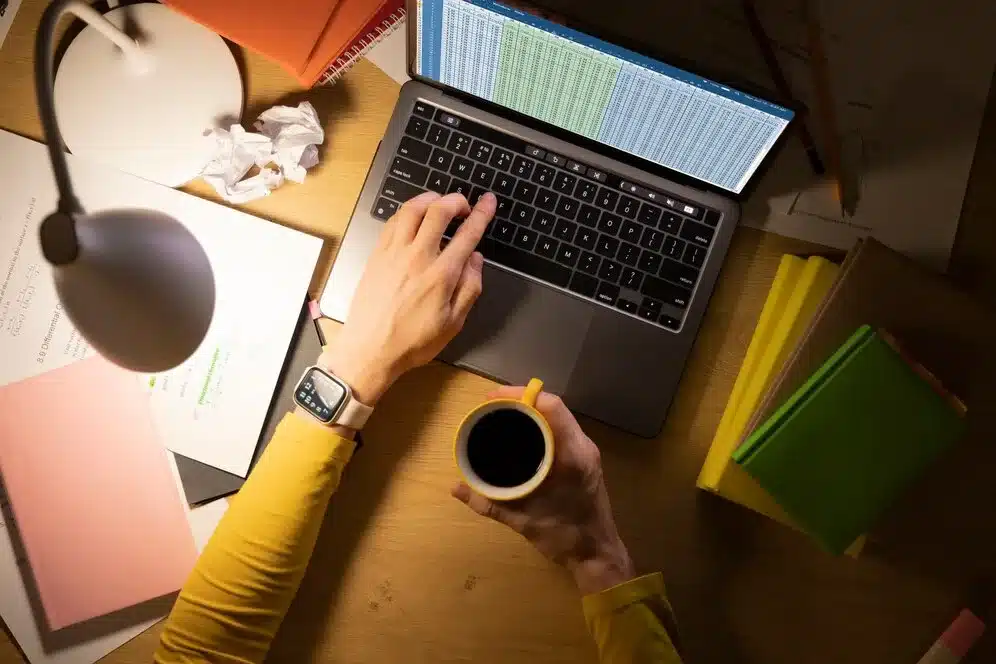 A imagem exibe um ambiente de trabalho com uma pessoa usando um notebook, com uma tela cheia de uma planilha eletrônica, provavelmente lidando com tarefas ou dados. A mão da pessoa está sobre o teclado, e ela segura um caneca de café, indicando um momento de pausa ou de concentração. No ambiente, há uma lampada, livros coloridos e papéis com anotações, sugerindo um ambiente organizado e produtivo.