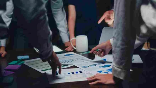 Grupo de profissionais em uma reunião de negócios, analisando gráficos e dados em uma mesa. Representa trabalho em equipe, planejamento estratégico e discussão de resultados. O ambiente é um escritório com uma mesa de madeira e um ambiente de trabalho colaborativo.