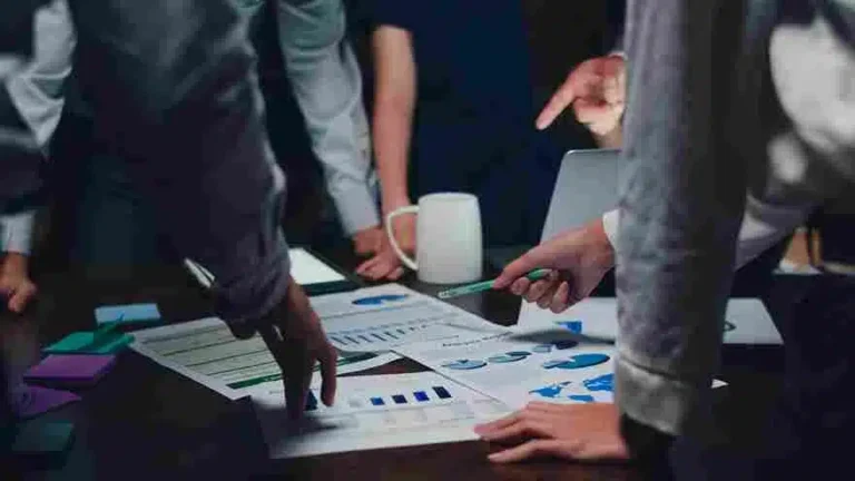 Grupo de profissionais em uma reunião de negócios, analisando gráficos e dados em uma mesa. Representa trabalho em equipe, planejamento estratégico e discussão de resultados. O ambiente é um escritório com uma mesa de madeira e um ambiente de trabalho colaborativo.
