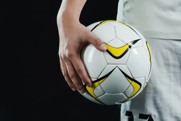 A imagem mostra uma mão segurando um bola de futebol, com detalhes em branco, preto e amarelo. A bola é um símbolo reconhecido do esporte, e a presença de uma pessoa com camisa de futebol indica que a imagem está relacionada a um contexto desportivo. No contexto do artigo "Como a Copa do Mundo vai impactar o varejo", a imagem pode ser usada para ilustrar o efeito que os eventos desportivos de grande escala, como a Copa do Mundo de Futebol, têm no varejo.