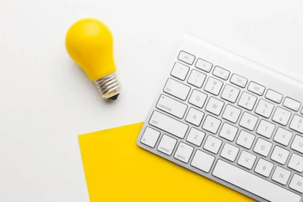 A imagem mostra um teclado de computador branco com uma lâmpada amarela em primeiro plano, sugerindo um ambiente de escritório ou local de trabalho. A imagem transmite a noção de estar preparado e ter estratégias definidas para o período pós-Black Friday, quando é necessário encantar e fidelizar os novos clientes adquiridos durante a campanha de vendas.