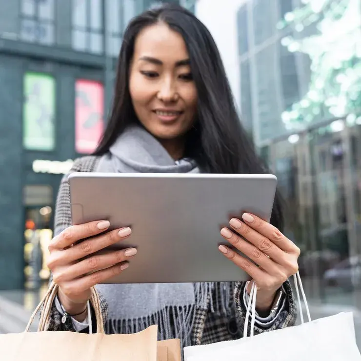 A imagem mostra uma mulher sorrindo e olhando para um tablet que ela está segurando com ambas as mãos. Ela está em uma rua, com sacolas de compras à sua volta, sugerindo que ela acabou de fazer compras ou está no meio de uma sessão de compras. O tablet está em uma posição que a cobre parcialmente, mas é claro que ela está interagindo com o dispositivo. No contexto do artigo "5 estratégias para o e-commerce em 2023: faça o seu negócio bombar", a imagem pode ser usada para ilustrar a experiência do consumidor no e-commerce. A mulher pode estar verificando promoções, comparando preços, ou gerenciando sua conta de compras online.