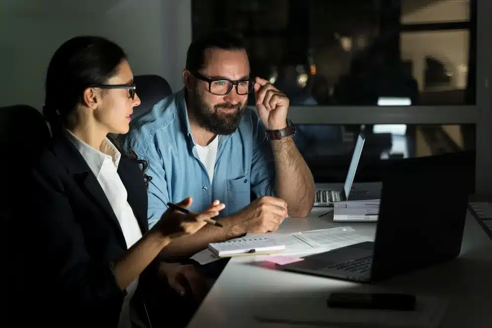 A imagem mostra um homem e uma mulher em um ambiente de trabalho, provavelmente em uma reunião ou discussão sobre negócios. O homem, que parece estar na posição de liderança ou consultor, está apontando para um documento e discutindo com a mulher, que está tomando notas. Ambos estão olhando para um laptop que está em frente a eles, o que indica que eles podem estar analisando dados ou informações relevantes ao ERP (Sistema de Planejamento Empresarial).