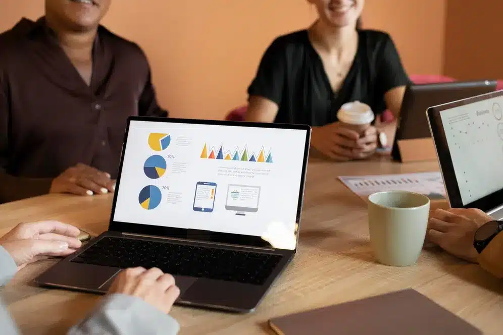 A imagem exibe uma reunião de trabalho onde três indivíduos estão envolvidos em atividades de análise de dados. O notebook no centro da imagem mostra gráficos de desempenho e estatísticas, o que sugere que os participantes estão examinando ou discutindo informações relevantes para o desempenho do negócio. O ambiente é informal, com um café à mão, indicando um ambiente de trabalho colaborativo e casual. No contexto do artigo "5 benefícios e recursos essenciais de um ERP para e-commerce", a imagem pode ser usada para ilustrar a importância de uma ferramenta de ERP (Sistema de Planejamento Empresarial) no gerenciamento e-commerce.