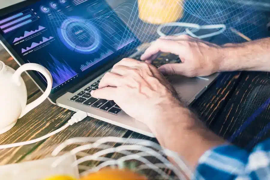 A imagem mostra uma pessoa usando um laptop com gráficos e dados na tela, indicando uma atividade de análise ou gerenciamento de informações. As mãos da pessoa estão sobre o teclado e o touchpad, sugerindo interação com o software. A tela do laptop exibe gráficos de linhas e um diagrama circular, o que pode representar relatórios de desempenho ou dados analíticos.