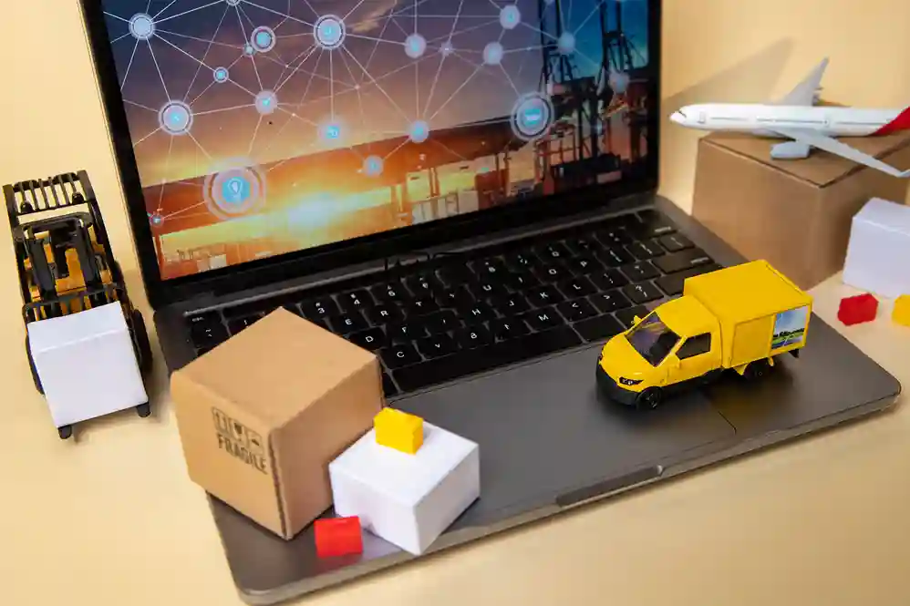 A imagem mostra a representação de uma cadeia de suprimentos digital e logística, com foco em tecnologia. Um laptop, com uma tela exibindo uma rede de conexões e ícones de transporte, serve como base. Sobre o teclado e ao lado do laptop, há miniaturas que simbolizam o fluxo de mercadorias: um caminhão de entrega amarelo, uma empilhadeira (forklift), e um avião de carga posicionado sobre uma caixa de papelão. As caixas de transporte (uma marrom com símbolos de "frágil" e uma branca) completam a cena. O conjunto ilustra a integração de logística, e-commerce, e transporte impulsionada pela tecnologia e pela gestão digital.