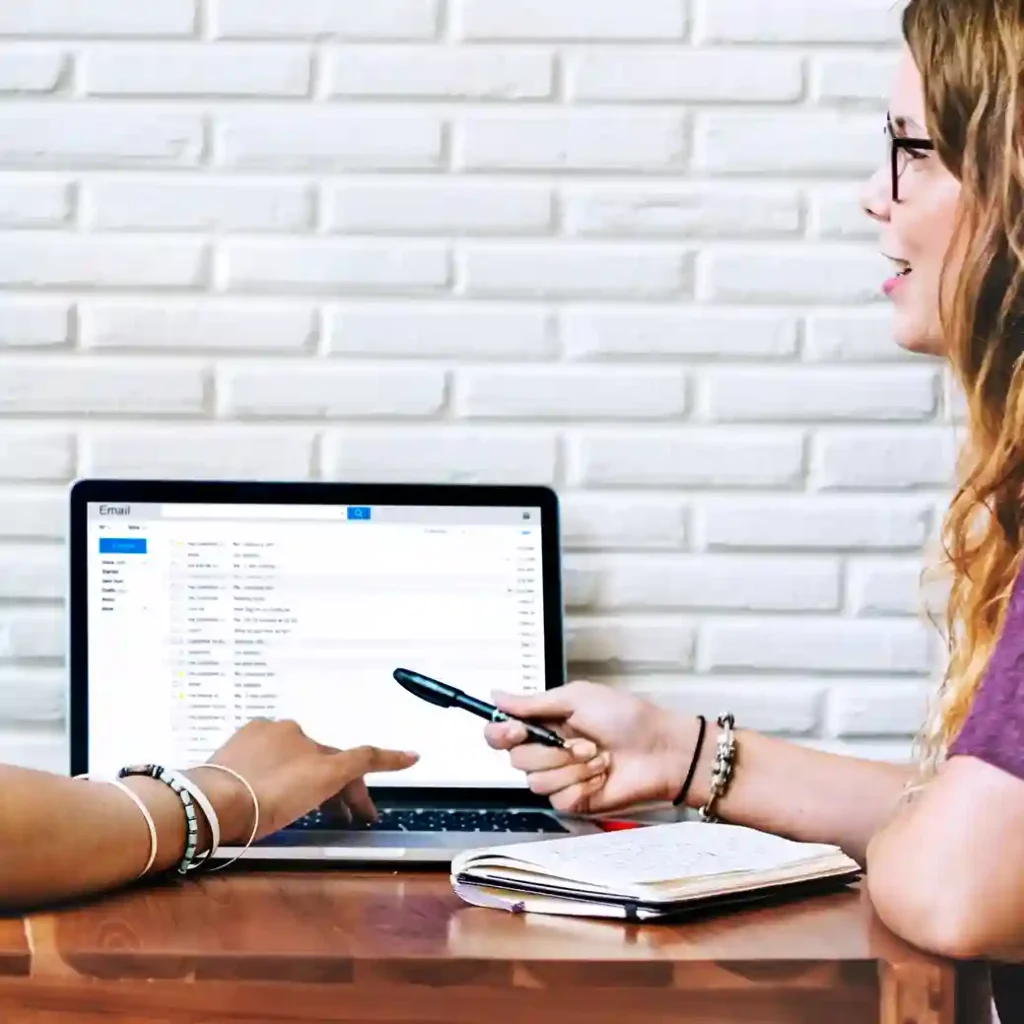 Duas mulheres em uma reunião de trabalho colaborativa em frente a um notebook. Uma delas aponta para a tela, que exibe uma interface de e-mail organizada, enquanto a outra observa. Sobre a mesa de madeira, há um caderno de anotações e uma caneta. A imagem ilustra o uso do Certificado Digital A1 para assinatura eletrônica de documentos, emissão de notas fiscais e autenticação segura em processos empresariais digitais.