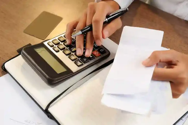 Close-up das mãos de um profissional utilizando uma calculadora financeira e uma caneta para conferir notas fiscais físicas sobre uma mesa de escritório. Ao lado, um caderno de anotações aberto. A imagem representa o processo de auditoria e a transição para um sistema de emissão de nota fiscal eletrônica automatizado, focando em precisão contábil e organização financeira.